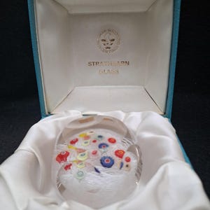 May include: A clear glass paperweight with a white, blue, red, and yellow floral design inside. The paperweight is in a blue box with a white interior and the words "Strathearn Glass" printed on the inside of the box.