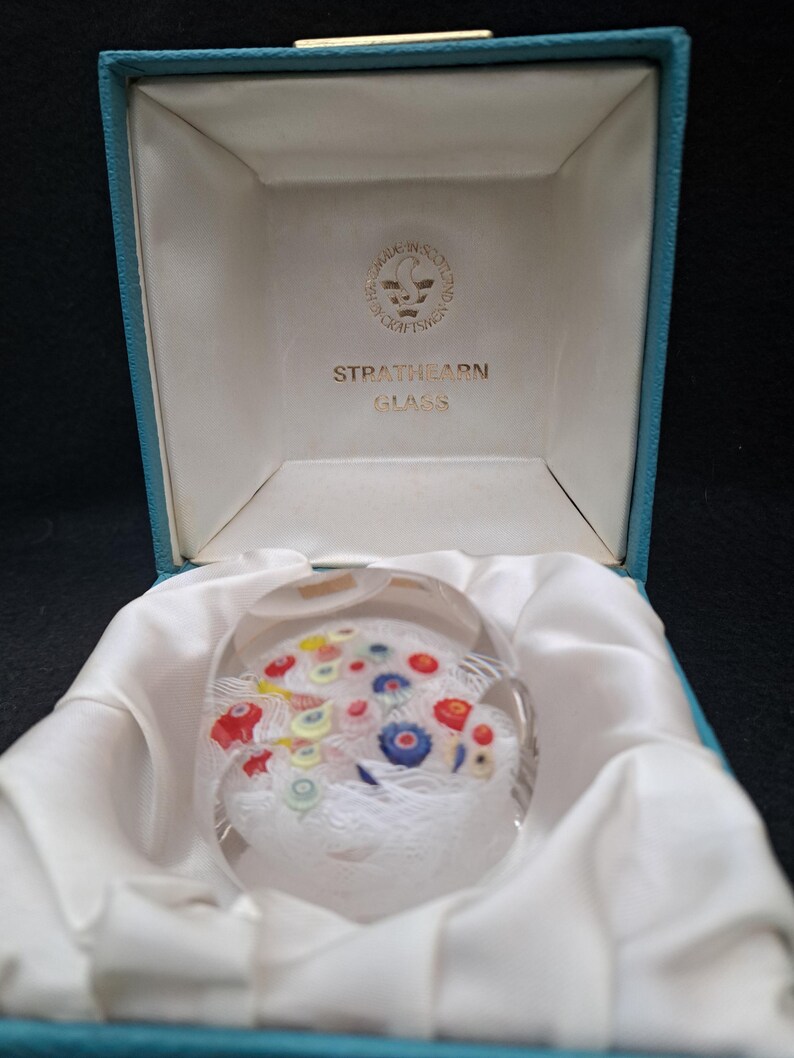 May include: A clear glass paperweight with a white, blue, red, and yellow floral design inside. The paperweight is in a blue box with a white interior and the words "Strathearn Glass" printed on the inside of the box.
