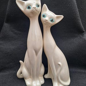 Op de afbeelding: Twee witte keramische kattenbeeldjes met blauwe ogen. De katten worden zittend afgebeeld, met slanke lichamen en puntige oren. Ze hebben een glanzende afwerking en een vintage esthetiek, in de stijl van modern design uit het midden van de eeuw.
