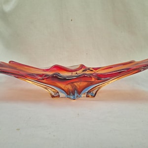 Op de afbeelding: Een decoratieve glazen schaal met een vloeiende, organische vorm. De schaal heeft een mix van kleuren, waaronder rood, oranje en amber, met hints van blauw aan de basis. Het ontwerp doet denken aan een bloem of een vlam.