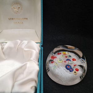 May include: A white glass paperweight with a colorful floral design inside. The paperweight is in a blue box with the words "Strathearn Glass" on the lid.