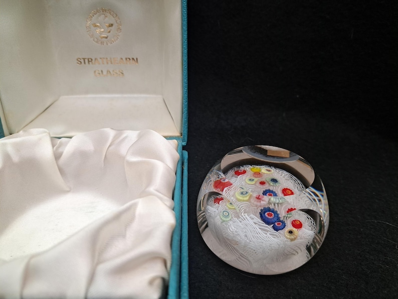May include: A white glass paperweight with a colorful floral design inside. The paperweight is in a blue box with the words "Strathearn Glass" on the lid.