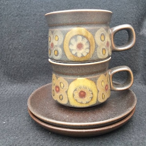 Könnte beinhalten: Zwei gestapelte braune Keramikbecher mit Henkeln und passenden Untertassen. Die Becher und Untertassen haben ein Blumenmuster in Gelb, Braun und Creme. Die Becher sind übereinander gestapelt, die Untertassen darunter.