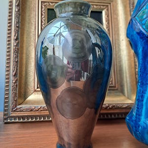 Può includere: Un vaso decorativo dalla forma bulbosa, con una sfumatura di colori dal marrone al blu. Il vaso ha una finitura lucida con disegni circolari marroni e grigi. Lo sfondo comprende un quadro con cornice dorata e una scultura in ceramica blu.