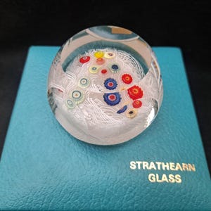 May include: A clear glass paperweight with white swirls and colorful flowers inside. The paperweight is sitting on a teal blue box with the words "Strathearn Glass" printed on it.