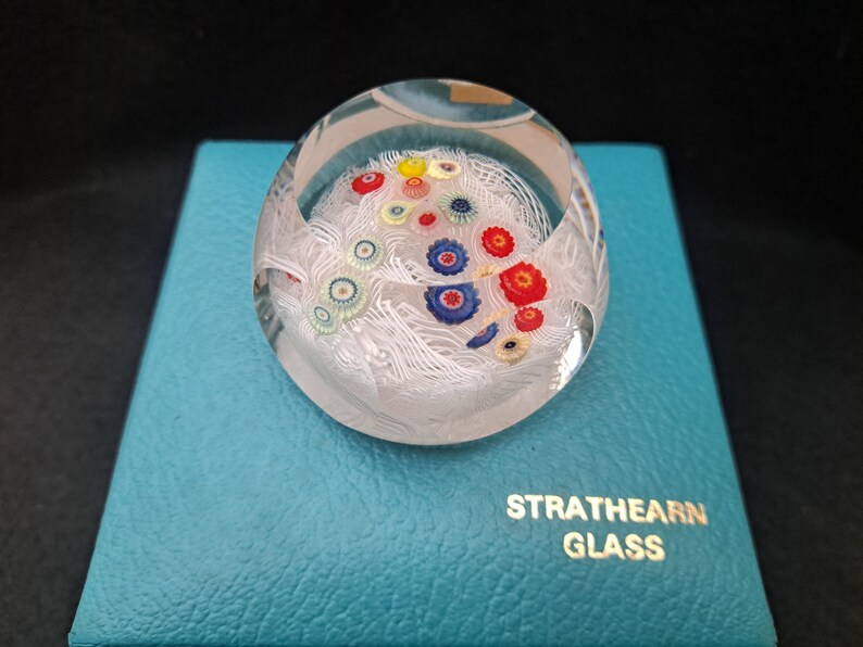 May include: A clear glass paperweight with white swirls and colorful flowers inside. The paperweight is sitting on a teal blue box with the words "Strathearn Glass" printed on it.