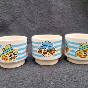 Puede incluir: Tres hueveras de cerámica blanca con rayas azules y blancas. Cada taza presenta un espantapájaros de dibujos animados con una expresión facial diferente.