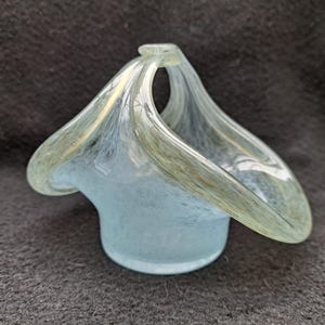 Op de afbeelding: Een kleine, mondgeblazen glazen mand met een handvat. De mand is lichtblauw met witte en gouden strepen. Het handvat buigt sierlijk vanaf de rand. De mand heeft een ronde basis en een uitlopende bovenkant.