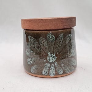 Peut inclure: Un pot en céramique avec un couvercle en bois. Le pot est brun foncé avec un grand motif floral dans les tons de bleu sarcelle et de blanc. Le couvercle est de couleur marron clair. Le pot est probablement utilisé pour le stockage.