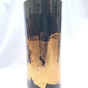 May include: A black glass vase with gold leaf accents. The vase is cylindrical in shape and has a smooth, glossy finish.