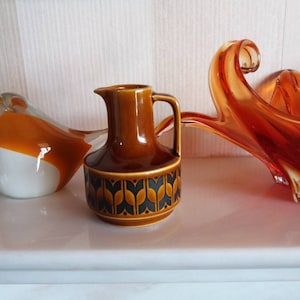 May include: A brown ceramic jug with a geometric pattern, a white and orange glass bird figurine, and a red glass bowl with a wavy design.