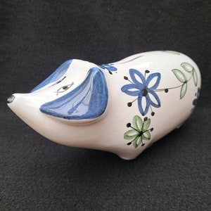 以下が含まれることがあります： 青と緑の花柄が描かれた白い陶器の貯金箱。貯金箱は青い鼻と耳をしています。