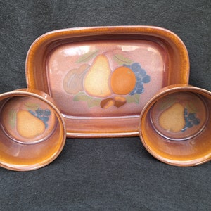 Può includere: Un set di tre piatti in ceramica marrone con un design a frutta. Il piatto grande ha una pera, un'arancia e dell'uva. I due piatti più piccoli hanno una pera e dell'uva.