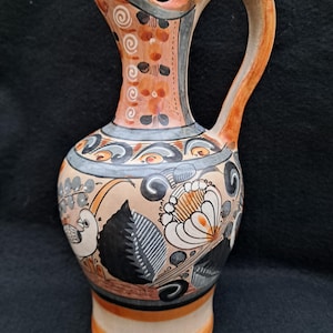 May include: Hand-painted ceramic pitcher with a curved handle and spout. The pitcher features a beige background with floral and bird designs in black, white, orange, and gray. The handle and rim are orange.