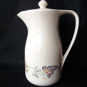 May include: White ceramic jug with a lid and a handle. The jug features a floral design in shades of blue, red, yellow, and green.