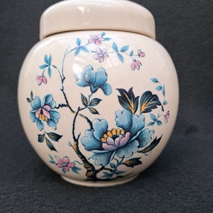 May include: A white ceramic jar with a lid, featuring a floral design in shades of blue, pink, and yellow. The flowers are stylized and have a vintage aesthetic.