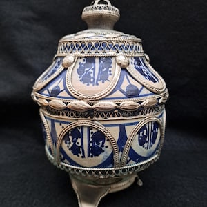 Può includere: Un vaso in ceramica con un motivo geometrico blu e bianco, impreziosito da bordi argentati e un coperchio argentato. Il vaso ha tre piedini argentati e una fascia argentata intorno alla parte superiore.