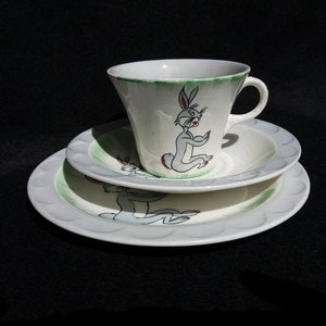 May include: A white teacup and saucer set with a green rim and a cartoon rabbit design. The rabbit is gray with red eyes and is sitting on a green patch of grass.