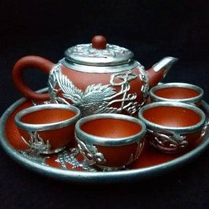 以下が含まれることがあります： 銀色の蓋と複雑な銀の模様が施された赤い銀の中国茶器セット。ティーポットと4つのカップが含まれています。カップにも銀の模様が施されています。