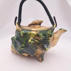 May include: A green and brown ceramic teapot shaped like a seashell with a black handle. The teapot has a textured surface and a spout on the side.