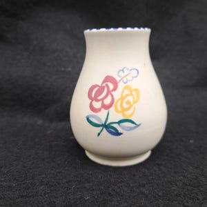 May include: A small, off-white ceramic vase with a flared rim and a rounded body. The vase is decorated with hand-painted flowers in pink, yellow, and blue. The rim has a blue trim.