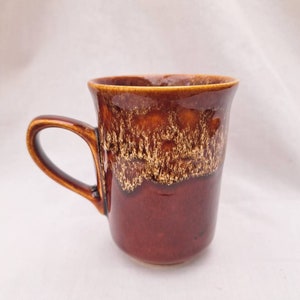 May include: A brown ceramic mug with a mottled glaze pattern. The mug has a rounded handle and a wide, slightly flared rim.