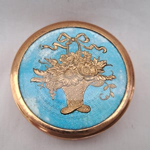 May include: A circular, antique compact with a gold-tone rim and a light blue enamel centre. The centre features a gold-tone relief of a flower basket with a bow. The compact is a vintage accessory.