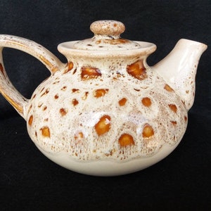May include: A white ceramic teapot with a brown speckled glaze. The teapot has a round body and a curved spout. The lid has a knob on top.