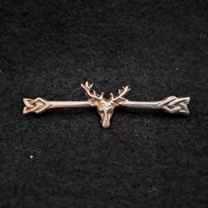 May include: A silver brooch with a deer head and antlers at the centre, with decorative arrow-like designs on either side. The brooch has a symmetrical design and is likely made of metal.
