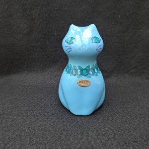 Puede incluir: Una figurita de cerámica de gato azul claro con ojos verdes y un diseño floral alrededor del cuello. La figurita tiene una etiqueta dorada que dice "DECO".