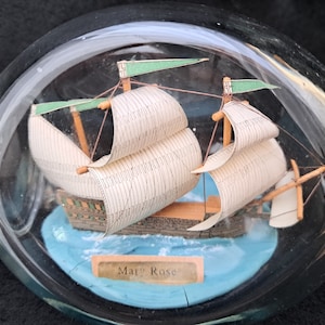 May include: A glass bottle containing a miniature model of the Mary Rose ship. The ship has white sails and green flags. The bottle has a blue base and a label that reads "Mary Rose."