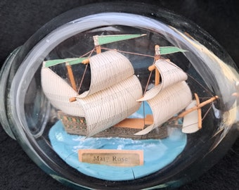 Op de afbeelding: Een glazen fles met een miniatuurmodel van het schip Mary Rose. Het schip heeft witte zeilen en groene vlaggen. De fles heeft een blauwe basis en een label met de tekst "Mary Rose."
