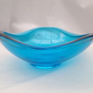 Puede incluir: Un cuenco de vidrio turquesa vibrante con un diseño curvo único. El cuenco tiene un acabado liso y brillante y un borde oscuro, creando un contraste llamativo. El cuenco probablemente se utiliza con fines decorativos.