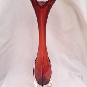 Puede incluir: Un jarrón de vidrio rojo alto y delgado con una abertura acampanada y una base cuadrada. El jarrón está hecho de vidrio transparente con un tinte rojo.