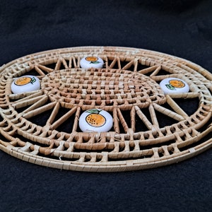 May include: A round woven placemat with a natural brown color and a decorative design. Four small white ceramic coasters with orange fruit designs are placed on the mat.