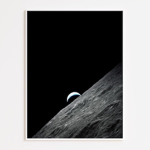 May include: A framed print featuring a black and white image of the moon's surface with the Earth rising in the background. The Earth is a blue and white sphere. The moon's surface is textured and gray.