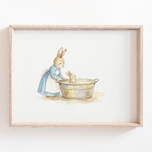 May include: A framed watercolour print depicting a mother rabbit in a blue dress and white apron, bathing a baby rabbit in a metal tub. The artwork is presented in a light wood frame, reminiscent of a classic children's story.