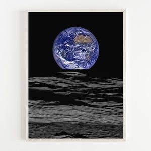 May include: A framed print featuring a vibrant image of Earth, showcasing blue oceans and landmasses, set against a black space backdrop. The planet appears to be rising above a textured, gray lunar surface, creating a striking contrast.