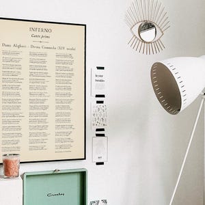 May include: A framed print of Dante Alighieri's Inferno, Canto Primo, hangs on a white wall. The text is in Italian. A white tripod floor lamp and a decorative eye-shaped mirror are also visible.