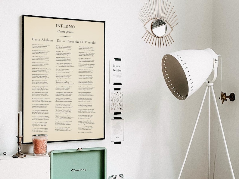 May include: A framed print of Dante Alighieri's Inferno, Canto Primo, hangs on a white wall. The text is in Italian. A white tripod floor lamp and a decorative eye-shaped mirror are also visible.