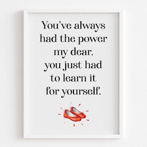 Op de afbeelding: Een wit ingelijst print met een citaat uit The Wizard of Oz: "Je hebt altijd de kracht gehad, schat, je moest het alleen leren om het zelf te doen." De quote staat in zwarte tekst en er zijn rode robijnenschoentjes onderaan de print.