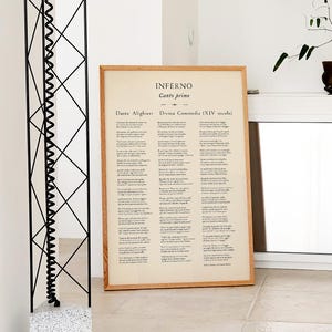 May include: A framed print of a page from Dante Alighieri's "Inferno" with text in Italian. The print has a light wood frame and is leaning against a white wall. A black metal sculpture and a potted plant are also visible.