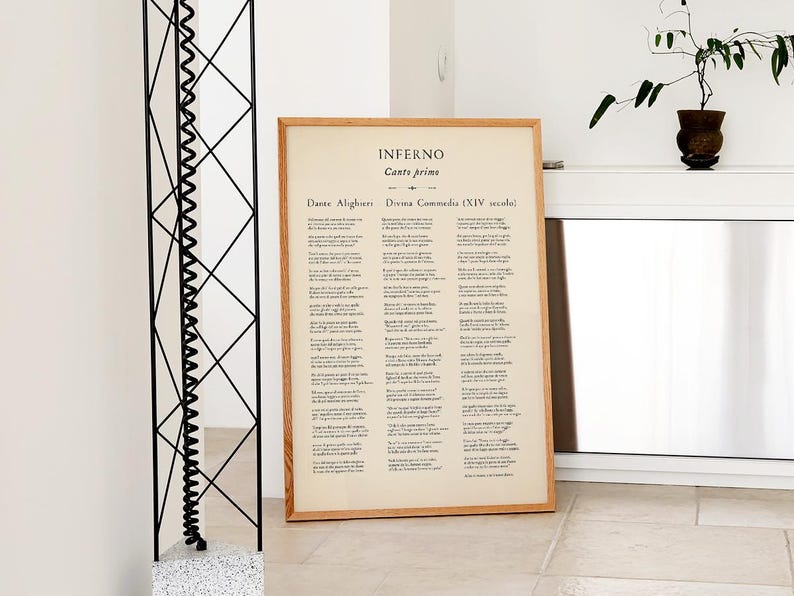 May include: A framed print of a page from Dante Alighieri's "Inferno" with text in Italian. The print has a light wood frame and is leaning against a white wall. A black metal sculpture and a potted plant are also visible.