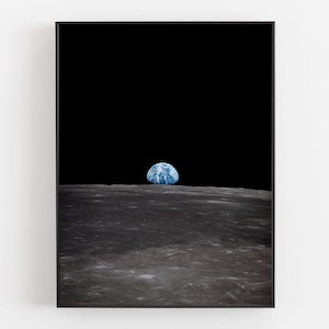 Può includere: Una stampa incorniciata raffigurante la Terra che sorge sopra la superficie lunare. La Terra è una sfera blu e bianca su sfondo nero. Il primo piano mostra la superficie grigia e strutturata della luna.
