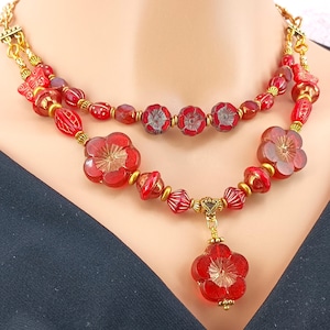 May include: A two-strand red glass bead necklace with a flower pendant. The necklace features gold accents and red glass beads in various shapes and sizes.