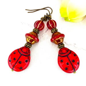 May include: A pair of dangle earrings featuring red ladybug-shaped beads with black spots. Each earring includes a bronze-toned metal hook, a small ribbed red bead, a larger metallic bronze spacer, and a textured bronze bead above the ladybug.