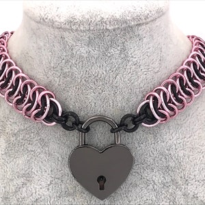 May include: A pink and black chainmail choker necklace with a heart-shaped padlock clasp. The necklace is made of interlocking metal rings, with pink rings on the outside and black rings on the inside. The heart-shaped padlock is gunmetal gray.