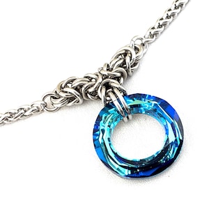 Peut inclure: Un collier argenté avec un pendentif en cristal bleu. Le pendentif est de forme circulaire avec un design à facettes, affichant des nuances de bleu. La chaîne a un motif tissé, et le pendentif est attaché avec une boucle en métal argenté.
