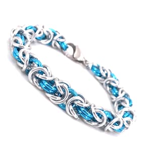 May include: A silver and blue chainmaille bracelet. The bracelet features a series of interlocking metal rings in silver and bright blue. The clasp is silver. The bracelet is shown on a white background.