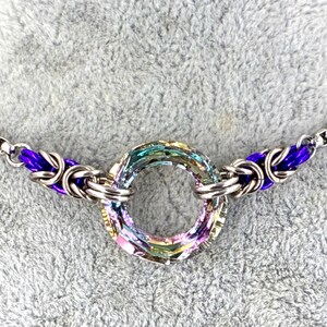 Peut inclure: Un collier argenté avec un pendentif central circulaire irisé. Le pendentif est entouré d'un motif de cotte de mailles avec des accents violets. La chaîne du collier est de type gourmette.